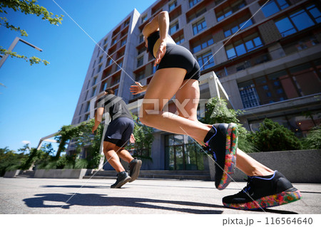 Low-angle view captures two runners sprinting in urban setting, with modern buildings towering above them. Fitness. 116564640