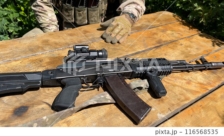 A close-up of a Kalashnikov assault rifle lies on the table in front of a soldier in uniform. 116568535