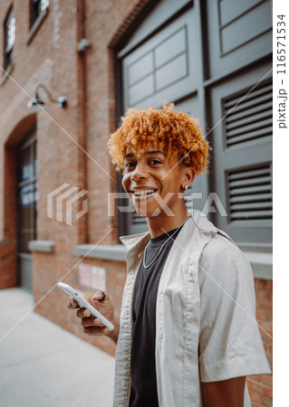 Smiling young man holding a smartphone in an urban environment with brick buildings 116571534