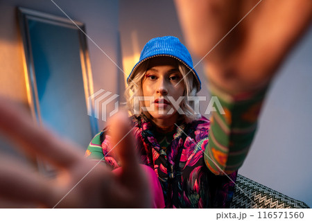 Fashionable individual in colorful attire with a blue hat reaching towards the camera 116571560
