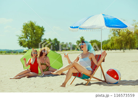 Women in stylish vintage swimsuits, have beach day. One relaxes in beach chair, other two lie under green parasol, all enjoying sunny weather. Women in stylish vintage swimsuits, have beach day. One relaxes in beach chair, other two lie under green parasol, all enjoying sunny weather. 116573559