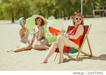 Group of women in vintage swimsuits, sipping drinks and soaking up sun on beach. One sits in beach chair, others lie on sand with parasol. Group of women in vintage swimsuits, sipping drinks and soaking up sun on beach. One sits in beach chair, others lie on sand with parasol. 116573561