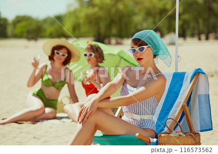 Three friends in retro swimwear, enjoying drinks and sunbathing on beach. One sits in chair under blue umbrella, while others relax with green parasol. Three friends in retro swimwear, enjoying drinks and sunbathing on beach. One sits in chair under blue umbrella, while others relax with green parasol. 116573562