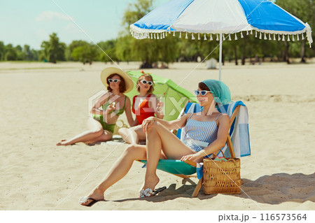 Three women in retro swimwear enjoying sunny beach day. One sits under umbrella with drink, while other two relax with green parasol. Vintage summer vibes. 116573564