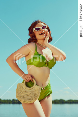 Joyful woman in red swimsuit and white cat-eye sunglasses, striking pose under sunny sky. Vintage summer charm. 116573582