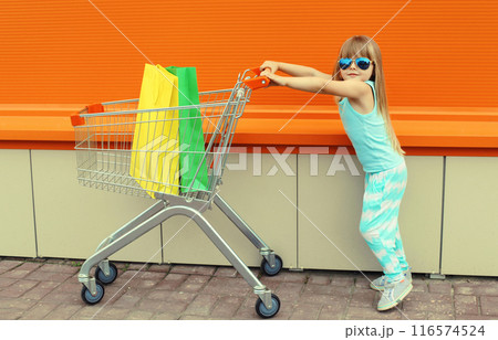 Happy little girl child walking with trolley cart and colorful shopping bags on city street 116574524