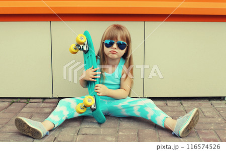 Stylish little girl child sitting on the ground with skateboard, happy kid posing on city street 116574526