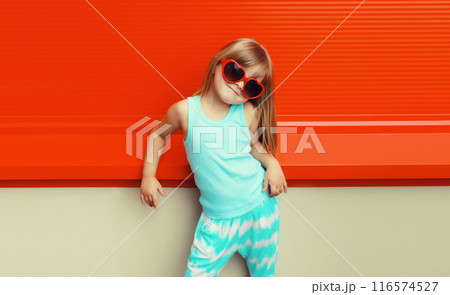Happy kid girl in red heart shaped glasses, stylish little child posing on city street on background 116574527