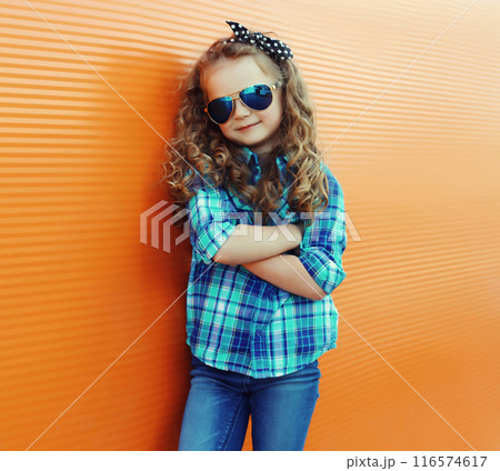Happy kid girl with curly hair, stylish little child posing in the city on orange wall 116574617
