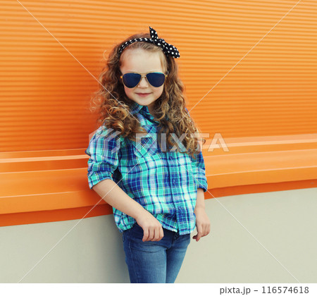 Happy kid girl with curly hair, stylish little child posing in the city on orange wall 116574618