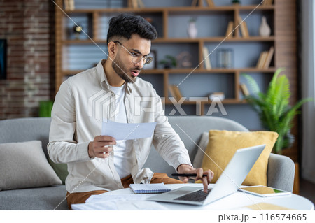 Focused young man working from home on couch analyzing financial documents using laptop. Remote work concept with home office setup displaying concentration and productivity 116574665