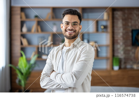 Portrait of a young Indian man wearing glasses and casual clothes, who is at home, standing with his arms crossed on his chest and smiling at the camera. 116574738