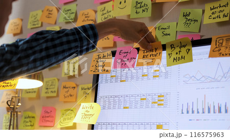 Male professional adds colorful notes to his computer screen amidst a vibrant array of papers, concept of managing time and planning 116575963