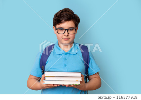 A cute schoolboy with backpack, in glasses and a blue T-shirt, holding a stack of books 116577919