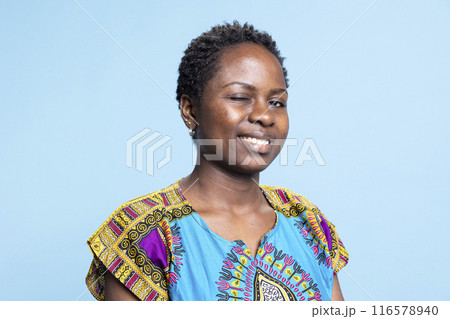 Portrait of african american girl winking and smiling on camera, feeling confident in her traditional authentic costume. Young serious woman posing over blue background, natural beauty. 116578940