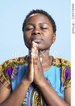 Confident serious person holding hands in a prayer to talk to God, saying grace and interacting with Jesus Christ in studio. Religious pious woman praying to the Lord, african american clothing. 116578948