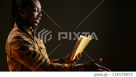 Young adult reading literature book on camera, standing over black background and enjoying lecture as hobby. African american person having fun with fiction or action novel story. Handheld shot. Young adult reading literature book on camera, standing over black background and enjoying lecture as hobby. African american person having fun with fiction or action novel story. Handheld shot. 116578953