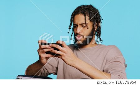 Man with disability in wheelchair playing videogames on cellphone, enjoying leisure time. Middle Eastern gamer enjoying game on mobile phone, having fun, studio background, camera B Man with disability in wheelchair playing videogames on cellphone, enjoying leisure time. Middle Eastern gamer enjoying game on mobile phone, having fun, studio background, camera B 116578954