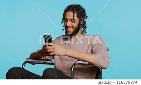 BIPOC man in wheelchair discussing with friend over online videocall using mobile phone. Person with paraplegia talking with mate in videoconference using cellphone, studio background, camera B BIPOC man in wheelchair discussing with friend over online videocall using mobile phone. Person with paraplegia talking with mate in videoconference using cellphone, studio background, camera B 116578974