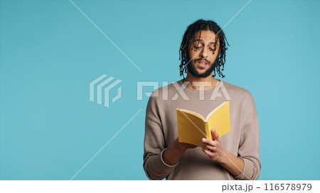 Middle Eastern man reading book from publisher aloud, telling story, enjoying hobby. BIPOC person narrating literature novel, doing critique essay, isolated over studio background, camera A Middle Eastern man reading book from publisher aloud, telling story, enjoying hobby. BIPOC person narrating literature novel, doing critique essay, isolated over studio background, camera A 116578979