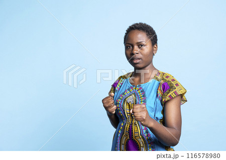 African american woman clenching fists and punching the air, showing strength and being aggressive in front of camera. Female model acting powerful and kickboxing, body training. African american woman clenching fists and punching the air, showing strength and being aggressive in front of camera. Female model acting powerful and kickboxing, body training. 116578980