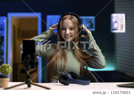 Joyous teenager filming herself enjoying new album from favorite musician, reviewing music in apartment. Merry content creator in home studio rating songs, hearing them through headphones 116579001