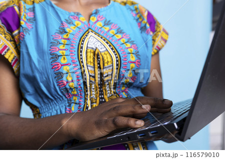 African american model navigating on internet webpages over blue background, using wireless computer. Young adult dressed in ethnic tribal clothing items uses laptop to check social media. 116579010