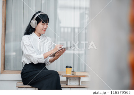 Young woman wearing headphones, using a tablet with a stylus, sitting outdoors near a coffee cup, looking focused and engaged. 116579563