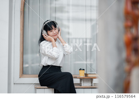 A young woman in a white shirt and black skirt enjoys music with headphones while sitting by a window with a coffee cup. 116579564