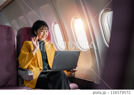 Professional woman in yellow blazer using laptop and waving while seated in airplane cabin, sunlight streaming through windows. Professional woman in yellow blazer using laptop and waving while seated in airplane cabin, sunlight streaming through windows. 116579572
