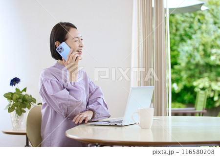リビングでスマホを操作しながらパソコンを見るミドル女性 116580208