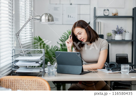 A young woman working from home, using a tablet and reviewing documents in a modern home office setting. A young woman working from home, using a tablet and reviewing documents in a modern home office setting. 116580435