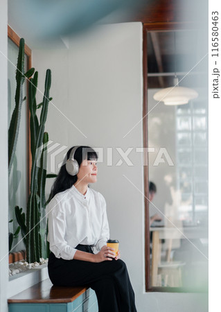 A young woman wearing headphones and holding a coffee cup, sitting in a modern minimalist cafe with cactus decor. A young woman wearing headphones and holding a coffee cup, sitting in a modern minimalist cafe with cactus decor. 116580463