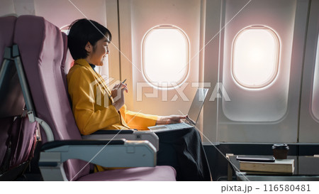 Professional woman in yellow jacket using laptop on airplane, working during flight, seated by window with coffee and book. Professional woman in yellow jacket using laptop on airplane, working during flight, seated by window with coffee and book. 116580481