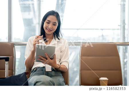 A charming Asian female passenger is working remotely, using her digital tablet in the airport. A charming Asian female passenger is working remotely, using her digital tablet in the airport. 116582353