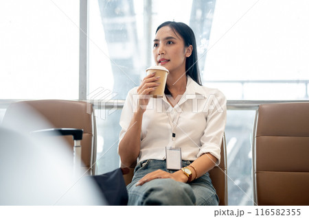 A confident Asian female passenger is having coffee while waiting for her flight in the airport. A confident Asian female passenger is having coffee while waiting for her flight in the airport. 116582355