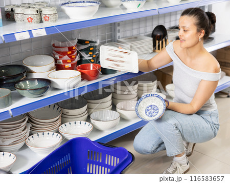 Positive Asian woman choosing plates in dishware store 116583657