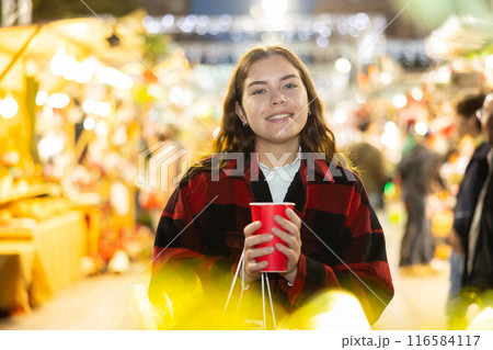Girl walking in colorful Christmas street market, holding paper cup of warm mulled wine 116584117
