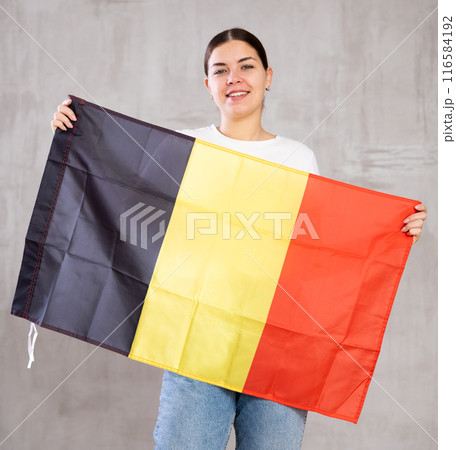 Glad young woman posing and smiling, standing with flag of Belgium against gray wall 116584192