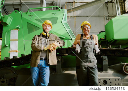 Medium long low angle view shot of African American woman and Caucasian man standing relaxed against industrial machine at work in factory Medium long low angle view shot of African American woman and Caucasian man standing relaxed against industrial machine at work in factory 116584558
