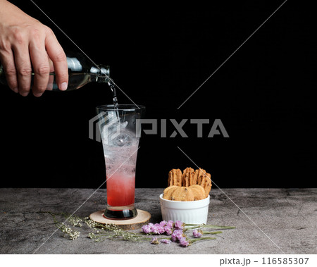 Hand pouring soda into glass with ice and red syrup on table with cookies and flowers, in dark background Hand pouring soda into glass with ice and red syrup on table with cookies and flowers, in dark background 116585307