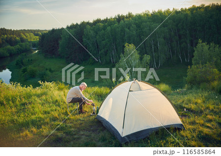 Extreme long shot of experienced senior male tourist sitting by camp tent cooking dinner, copy space 116585864