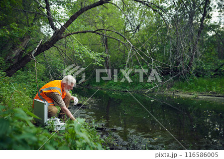 Wide shot of senior Caucasian male ecologist in safety vest taking samples at bank of river in forest on summer day, copy space Wide shot of senior Caucasian male ecologist in safety vest taking samples at bank of river in forest on summer day, copy space 116586005