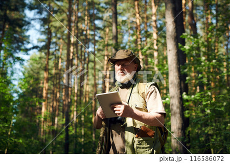 Medium shot of senior Caucasian male researcher standing in forest on sunny summer day and using digital tablet Medium shot of senior Caucasian male researcher standing in forest on sunny summer day and using digital tablet 116586072