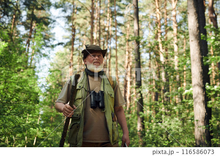 Medium shot of senior Caucasian man with beard on face wearing hat walking in forest with use of wooden stick, copy space Medium shot of senior Caucasian man with beard on face wearing hat walking in forest with use of wooden stick, copy space 116586073