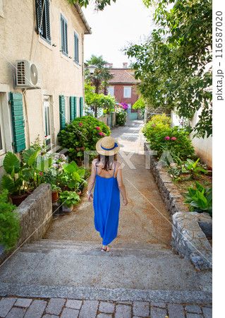 A woman in a blue dress and straw hat walks down a quaint alley lined with green plants and flowers. A woman in a blue dress and straw hat walks down a quaint alley lined with green plants and flowers. 116587020