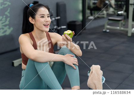 Happy young beautiful woman taking a break to eat her green apple during her morning work out at a local gym 116588634