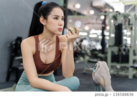 Happy young beautiful woman taking a break to eat her green apple during her morning work out at a local gym 116588636