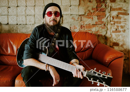 Mature Caucasian rock musician wearing stylish red sunglasses sitting relaxed on leather couch holding guitar and looking at camera, copy space Mature Caucasian rock musician wearing stylish red sunglasses sitting relaxed on leather couch holding guitar and looking at camera, copy space 116588677