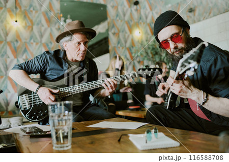 Two gen X Caucasian male rock musicians sitting at old wooden coffee table playing guitars, medium shot Two gen X Caucasian male rock musicians sitting at old wooden coffee table playing guitars, medium shot 116588708